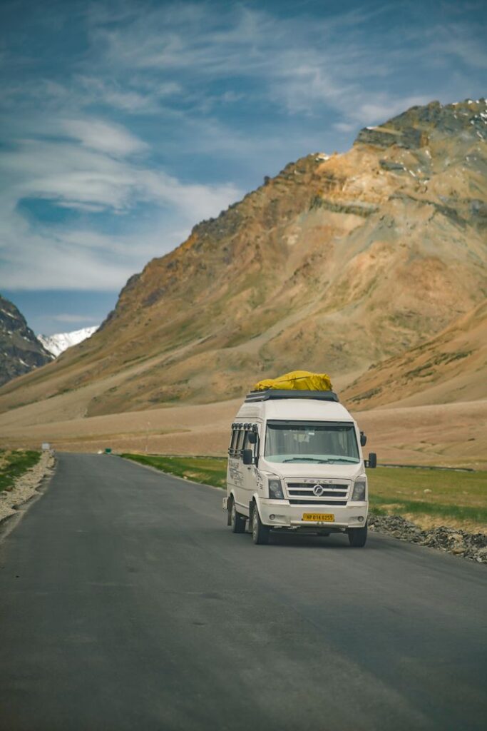 White van travels on a scenic road through the mountains of Leh, India, under a clear blue sky.